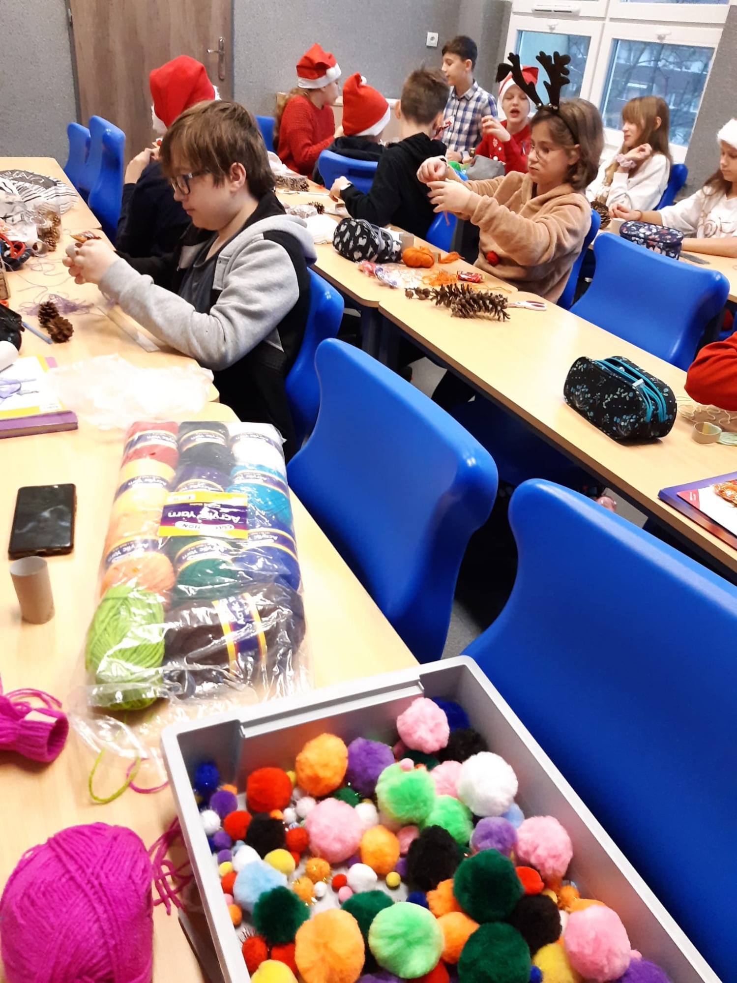 W klasie dzieci pracują przy ławkach w świątecznych czapkach. Na pierwszym planie na stole leżą kolorowe włóczki i pompony.