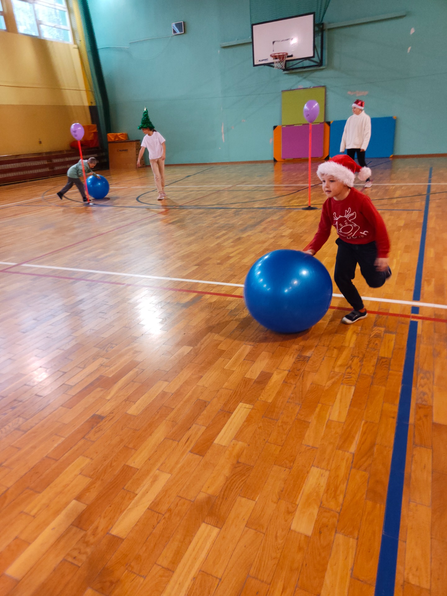 Dwójka dzieci w czapce Mikołaja toczy dużą niebieską piłkę.