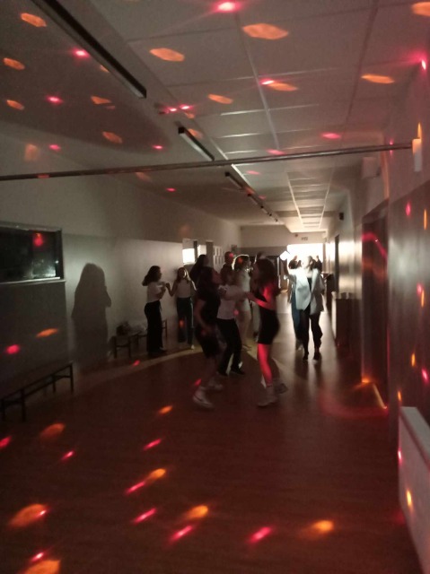 Na ciemnym tle korytarza szkolnego znajdują się tańczący uczniowie. W tle widać kolorowe światełka w odcieniach czerwieni.  .

