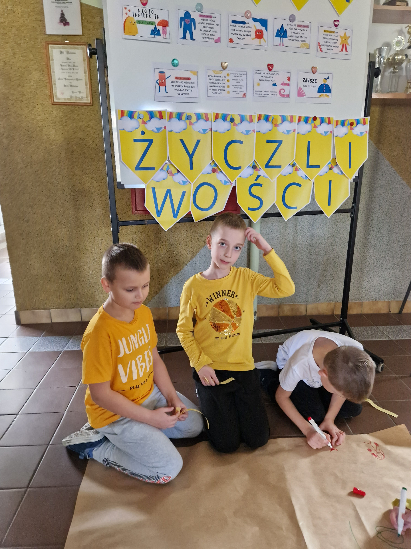 Trzej uczniowie siedzący  pod tablicą  z dekoracją, zamierzają  narysować  stworka życzliwości 