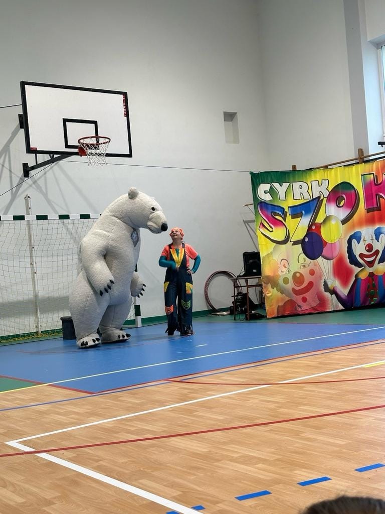 Zdjęcie przedstawia clowna stojącego na sali gimnastycznej. Obok niego stoi osoba przebrana za niedźwiedzia.