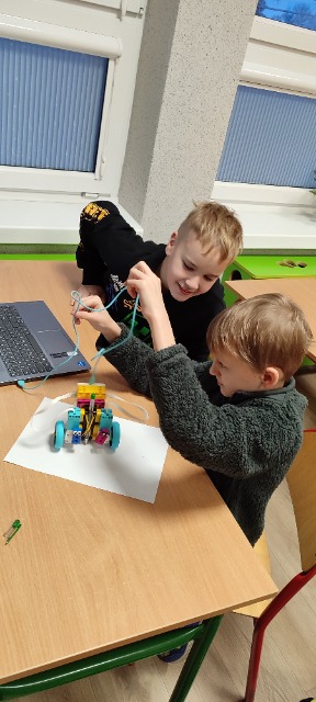  Dwóch uczniów siedzi przy stoliku szkolnym, na którym znajduje się laptop i zbudowany z klocków robot. Jeden z uczniów trzyma w ręce kabel.