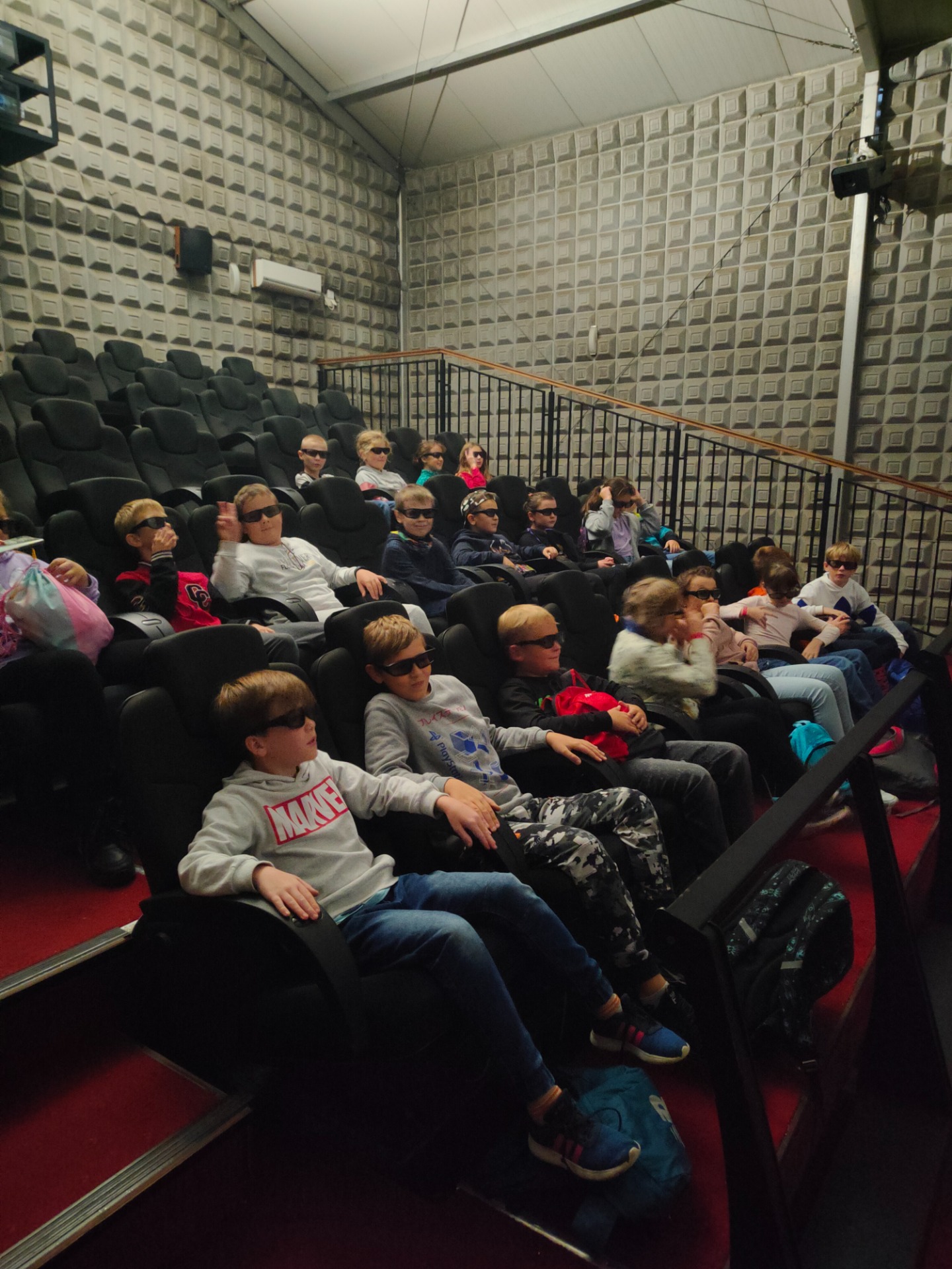 Grupa dzieci siedzą w kinie 5d. Wszyscy mają założone czarne okulary.