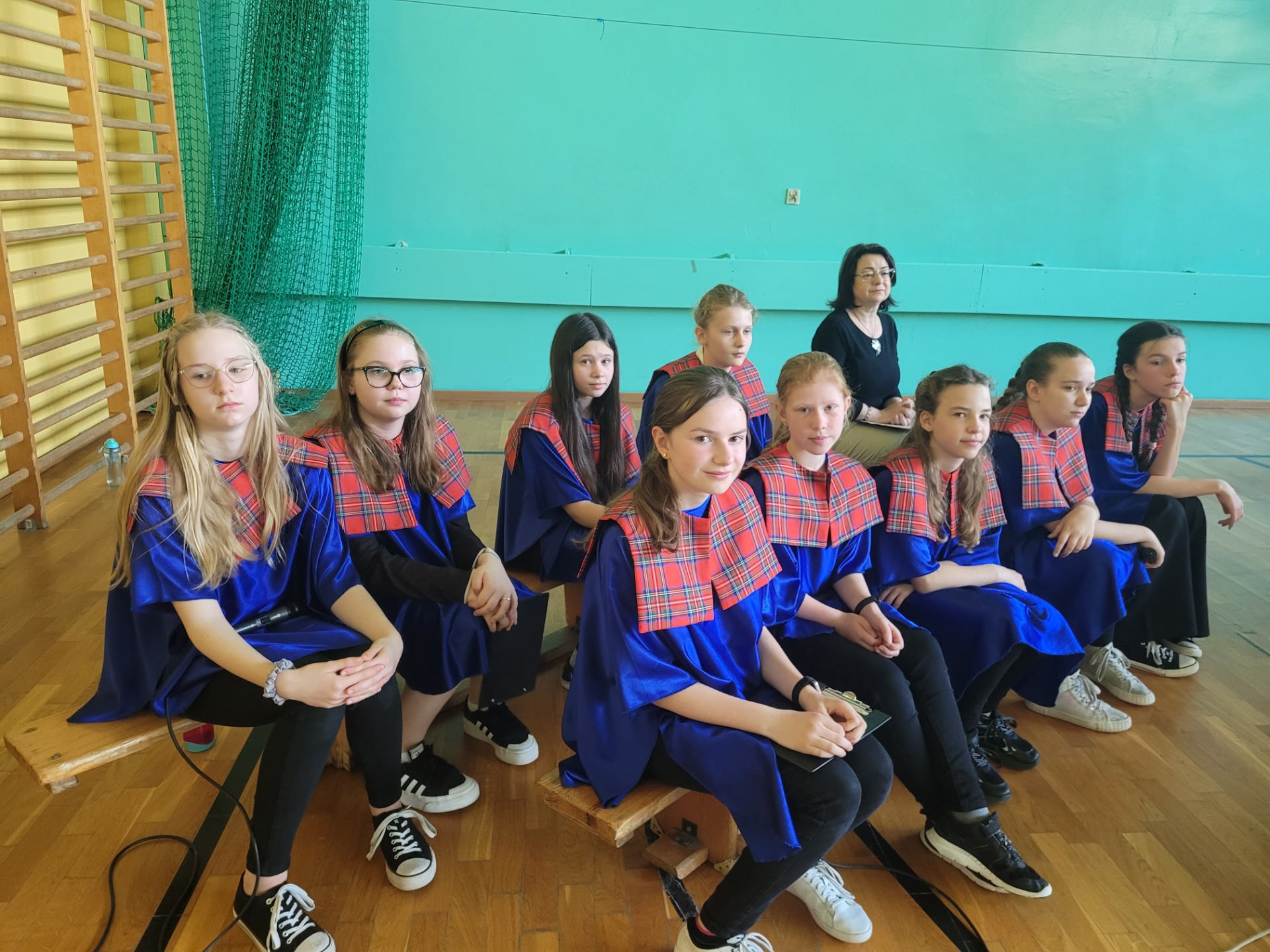 9 uczennic w strojach chóru (niebieskie bluzki z czerwonymi kołnierzami) siedzi na ławeczkach z nauczycielką muzyki.