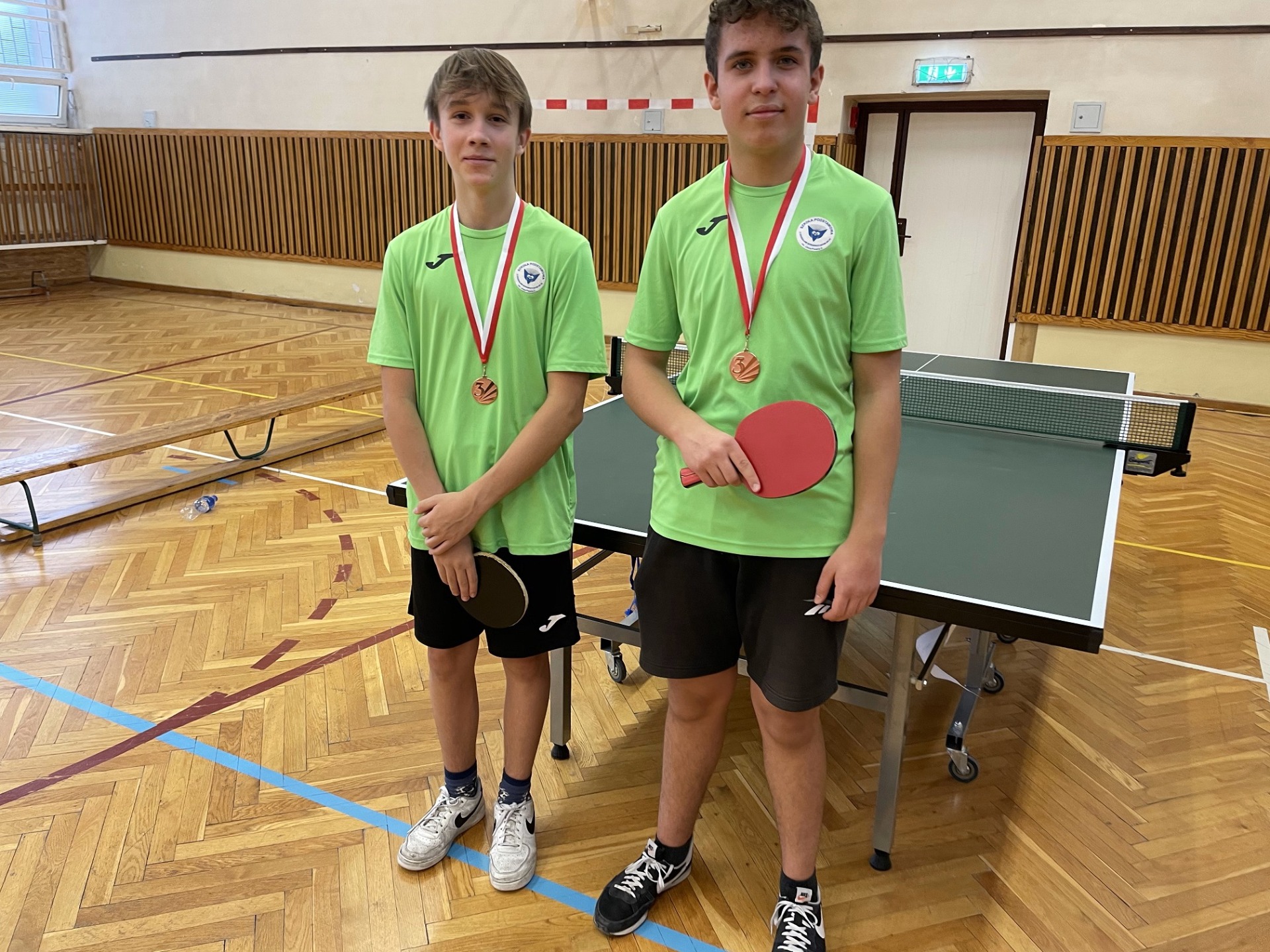 Zdjęcie przedstawia reprezentację chłopców gr. starsza, którzy ubrani są w zielone koszulki. W rękach trzymają paletki do tenisa stołowego a na szyi mają zawieszone medale.