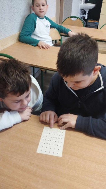 Dwóch uczniów siedzi przy ławce, pochylają się nad tabliczką z alfabetem Braille'a.