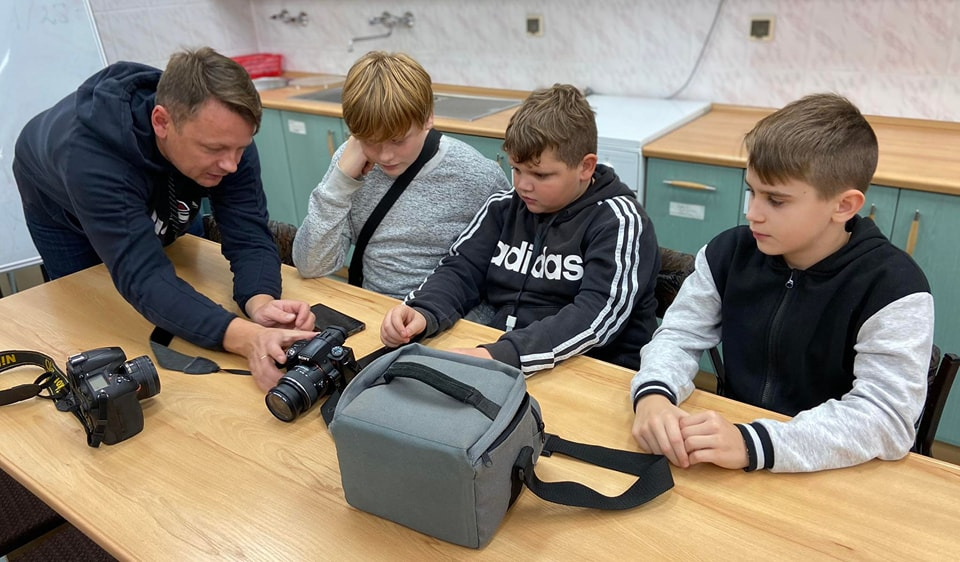 Warsztaty fotograficzne. Fotograf prezentuje sprzęt trojgu chłopcom.