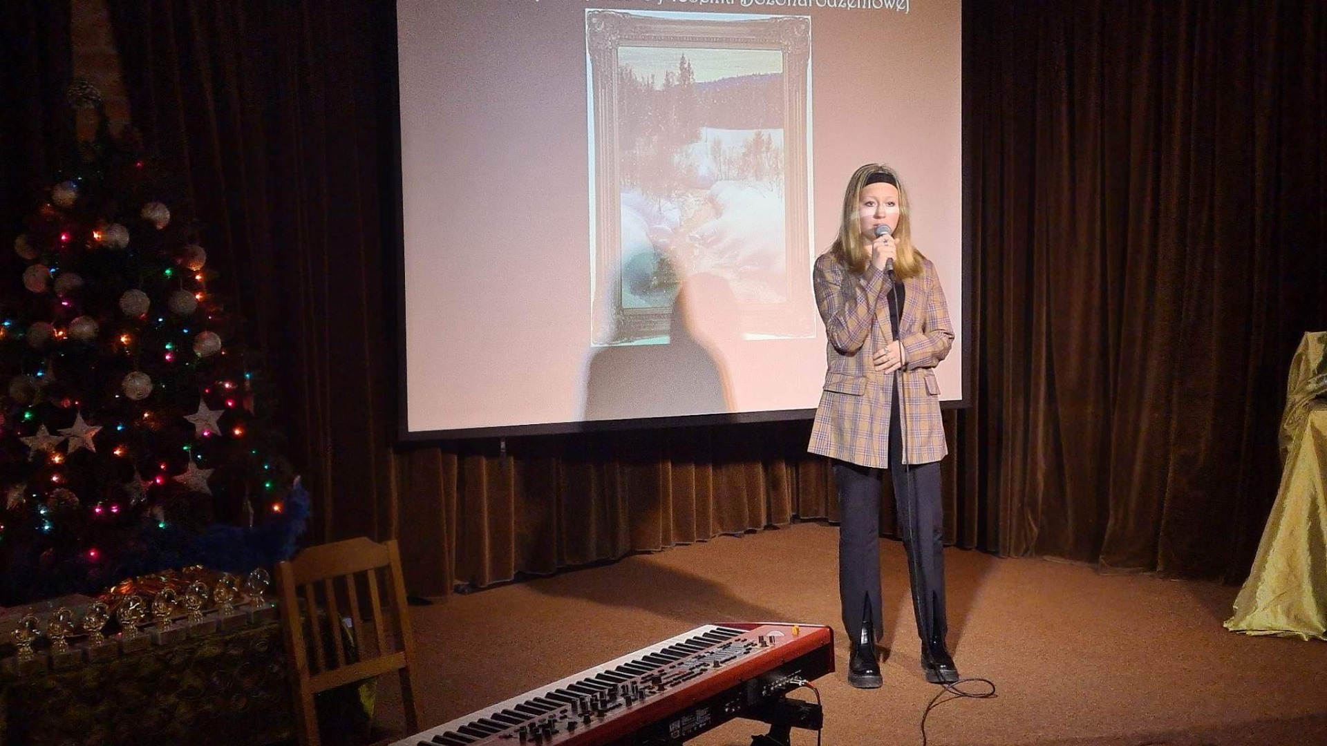 Dziewczyna w marynarce w kratkę i czarnych spodniach stoi na scenie. W ręku trzyma mikrofon. W tle widoczna dekoracja: prezentacja z motywami świątecznymi, choinka, pianino.