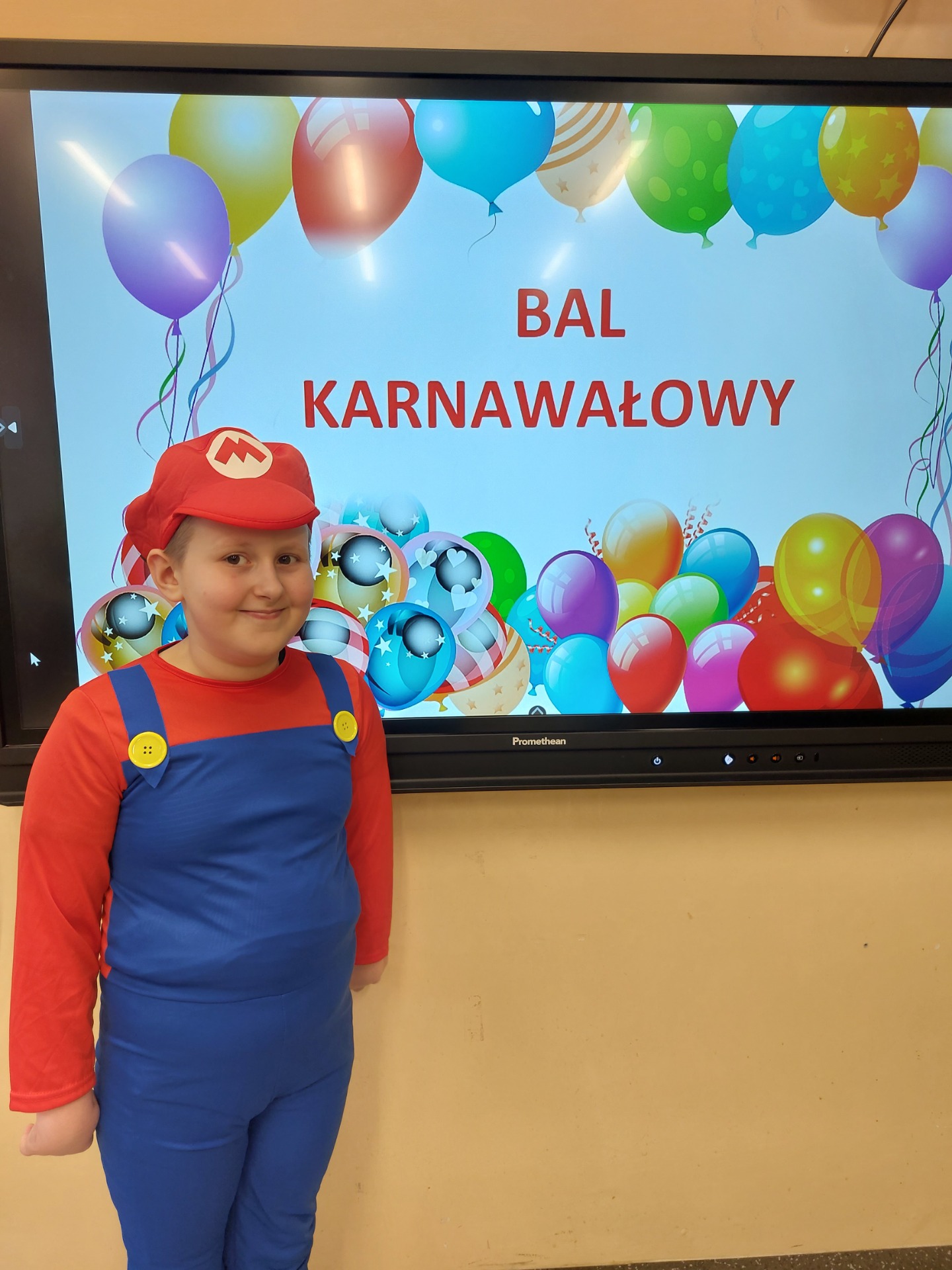 Uczeń przebrany za postać Mario pozuje do zdjęcia, z tyłu napis: Bal karnawałowy.