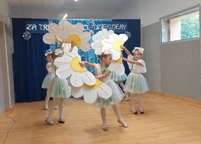 5 uczennic w biało-błękitnych strojach tańczy z biało-żółtymi kwiatami z tektury w rękach.