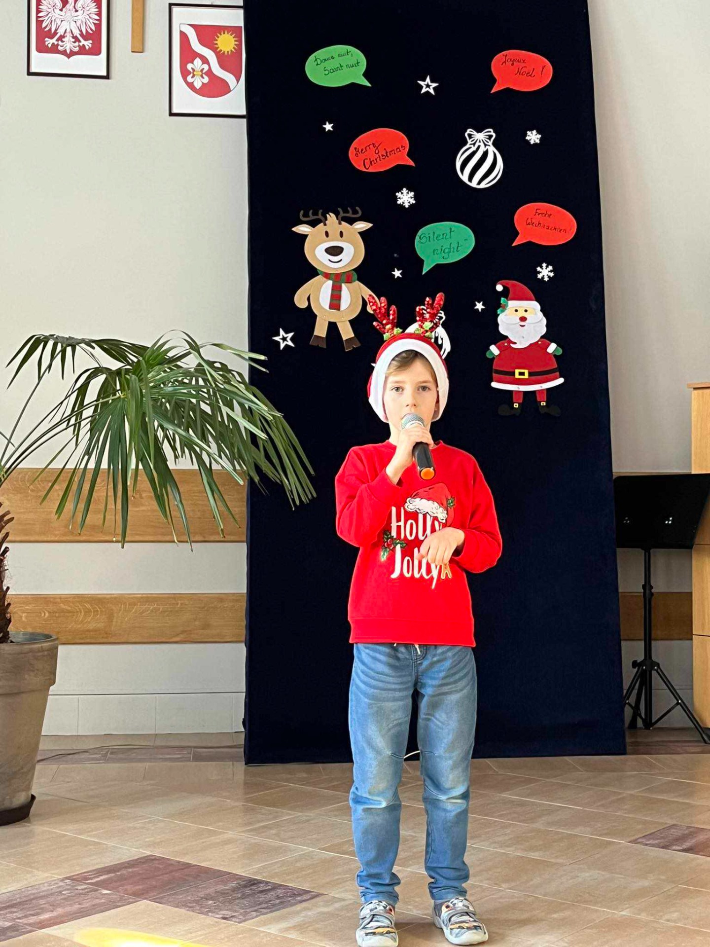 Na tle świątecznej dekoracji stoi chłopiec w czerwonym swetrze i czapce Mikołaja na głowie. Śpiewa do mikrofonu.