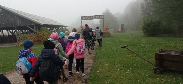 Grupa uczniów idzie w parach. Przed nimi dwie nauczycielki ,na wprost napis Wioska Dyniowa. Po prawej stronie stoi drewniany wózek , po lewej duża altanka.